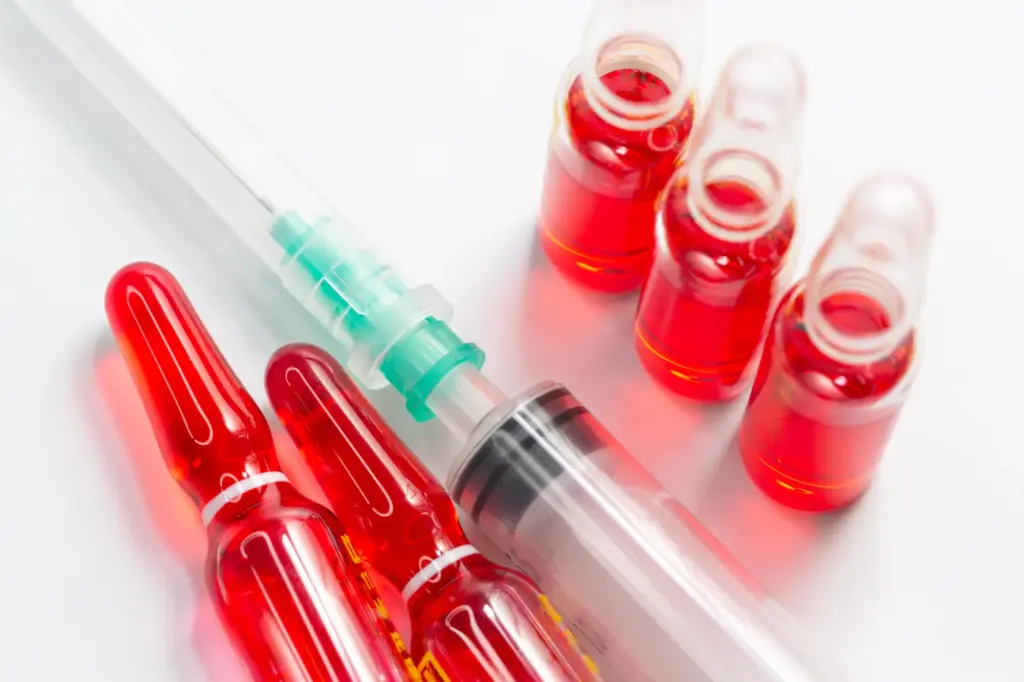 Close-up of a syringe and several glass vials filled with red liquid, arranged on a white surface, representing medical injections or vitamin treatments.
