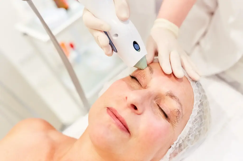Close-up of a woman receiving a non-invasive eye treatment, with a practitioner using a handheld device on the forehead area while wearing protective gloves in a clinical setting.