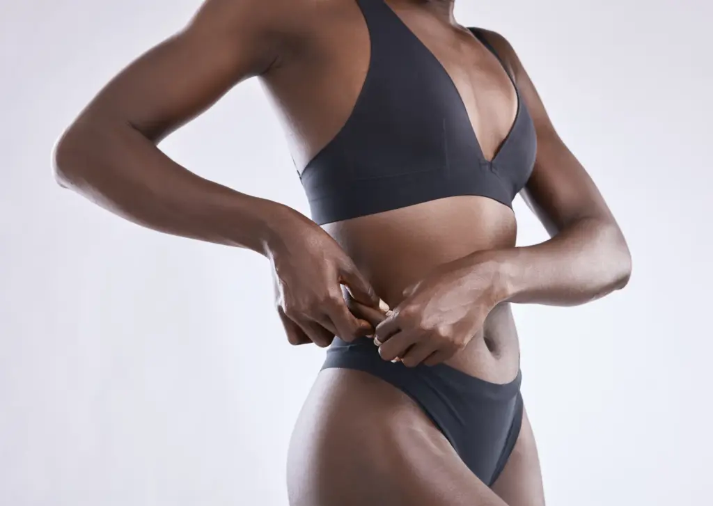 Close-up of a fit woman’s torso in black athletic underwear, adjusting the waistband at her hip, highlighting a toned abdomen and waist against a neutral background.